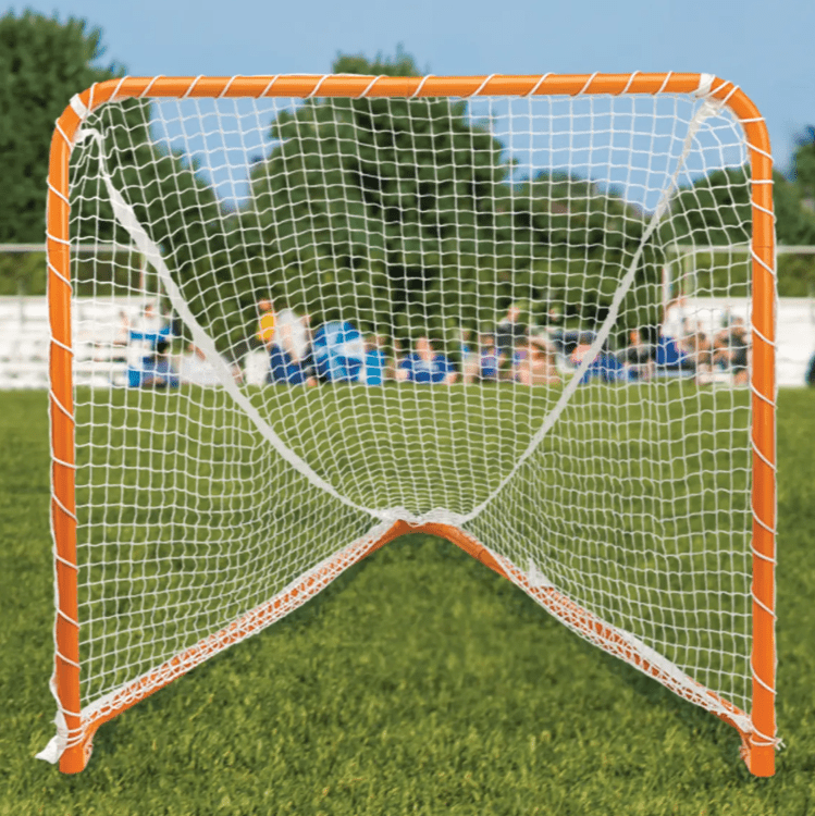A close-up view of an orange STX Folding Lacrosse Goal 4x4 with a white net on a green grass field. In the background, there are blurred figures of people sitting and white fencing, with trees and a blue sky overhead—a testament to the quality control of STX in outdoor sports gear.