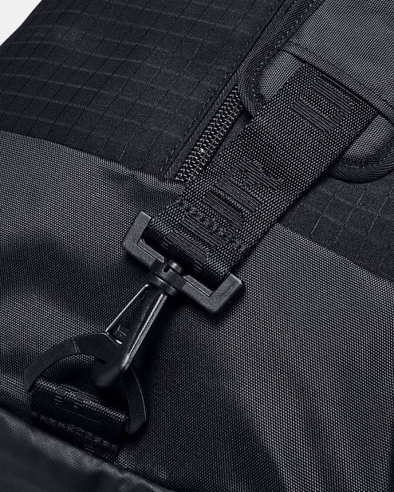 Close-up of the UA Lacrosse Equipment Bag by Under Armour featuring a textured strap with a plastic clip. The strap is connected to a grid-patterned section, and the clip is secured to a loop below. The materials look durable and rugged.