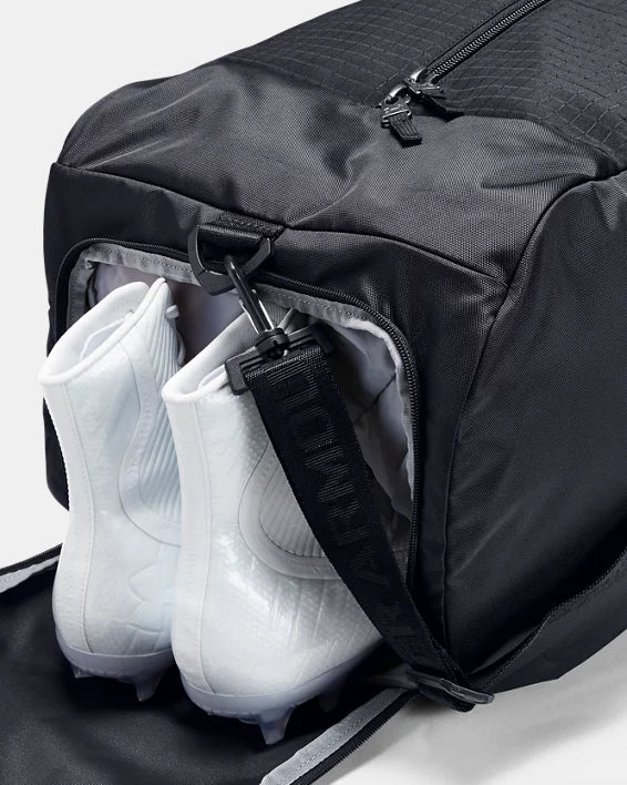 A black Under Armour 1352119 sports bag with an open side compartment displays a pair of partially inserted white soccer cleats. The bag features a black strap with a clip attached.